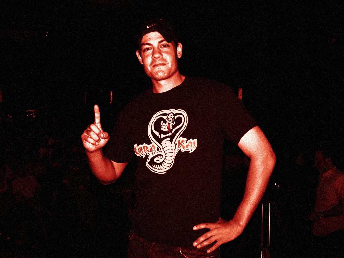 Alex Wolfe on stage in a Cobra Kai shirt, one finger raised